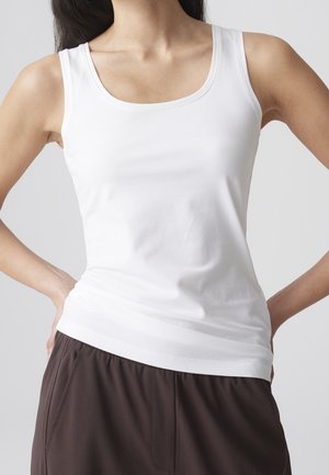 Femme portant un débardeur blanc uni sans manches et un pantalon foncé et ample, les mains posées sur les hanches sur un fond uni.