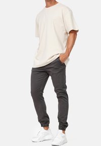 Vaaleanbeige ylisuurennettu t-paita, joka on yhdistetty tummanharmaisiin slim-fit-jogger-housuihin. Valkoiset lenkkarit täydentävät asua ja korostavat rentoa, nykyaikaista tyyliä.