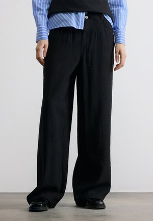 Osoba oblečená v voľných čiernych nohaviciach, čiernych topánkach a modrej pruhovanej košeli stojaca na sivom podlahe proti jednému bielemu pozadiu.