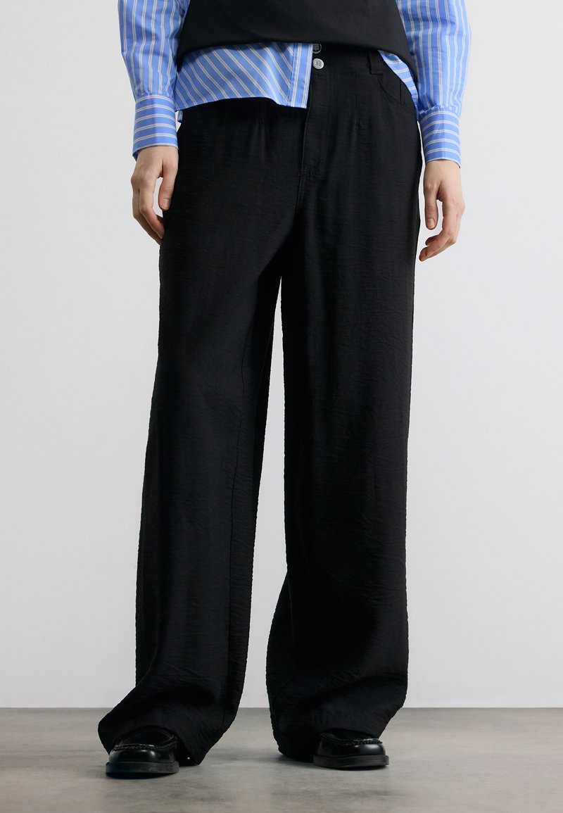 Personne portant un pantalon noir ample, des chaussures noires et une chemise rayée bleue, debout sur un sol gris devant un fond blanc uni.