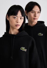 Los sudaderas con capucha negras presentan un logo de cocodrilo verde en el lado izquierdo del pecho, con cordones y una textura de tela suave. Dos modelos las llevan puestas.