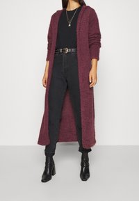 Cardigan lungo color bordeaux in un tessuto morbido, design aperto davanti, indossato sopra un top nero e jeans neri, abbinato a stivali neri con tacco.