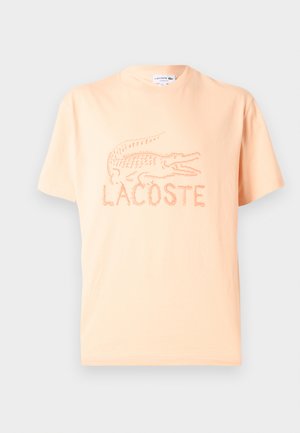 Κοντομάνικο T-shirt σε ανοιχτό ροδακινί χρώμα με ανάγλυφο υφασμάτινο λογότυπο κροκοδείλου Lacoste και το όνομα της μάρκας στο στήθος.