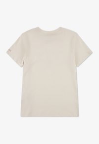 Beige bomullst-shirt med korta ärmar och rund halsringning. Har en liten rosa logotyp på vänster ärm. Slät textur, enkel design.