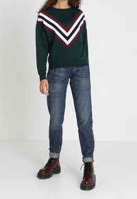 Sudadera verde con un gran diseño en forma de V en burdeos, blanco y navy, combinada con jeans de un azul oscuro y botas de cordones burdeos.