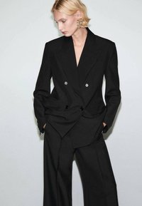 Czarna marynarka dwurzędowa z klapami szpiczastymi, dopasowanym krojem, bocznymi kieszeniami oraz szerokimi spodniami o prostych nogawkach w tym samym kolorze. Gładka, strukturalna tkanina.