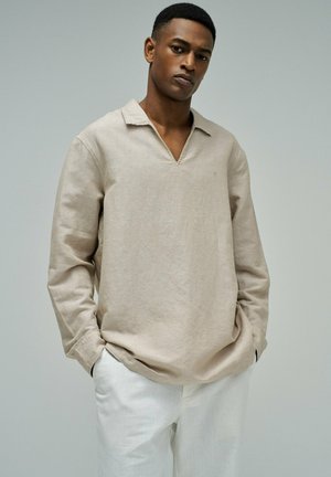 Homme les mains dans les poches portant une chemise beige à manches longues avec col et un pantalon blanc, devant un fond gris uni.