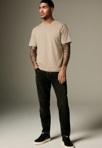 Beige bomulds t-shirt med korte ærmer, parret med mørke jeans og sorte sneakers. Personen har synlige tatoveringer på deres arme.