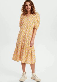 Vestido midi floral com mangas bufantes, base amarela e flores rosa-laranja. Apresenta fecho de botões na frente e barra em camadas. Combinado com botas bege.