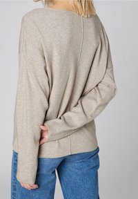Beige stickad tröja med sänkt axel, ett V-format ryggsöm och långa ärmar. Kombinerad med blå högmidjade jeans som har en avslappnad passform.