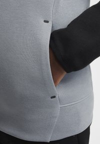 Sudadera gris y negra con una tela texturizada, bolsillos laterales y detalles de costura visibles, además de herrajes negros de acento.
