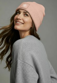 Knitted pink beanie with a ribbed texture and a square patch featuring a compass design. Worn with a gray sweater, hair flowing to one side.