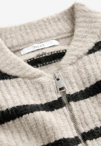 Beige stickad tröja med svarta ränder, med dragkedja. Texturen är mjuk och fluffig, med en etikett som säger "NEXT" inuti kragen.