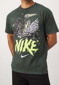 Camiseta de algodón verde con un atrevido gráfico en negro y amarillo de un jinete alado y el gran texto "NIKE". Mangas cortas, cuello redondo.