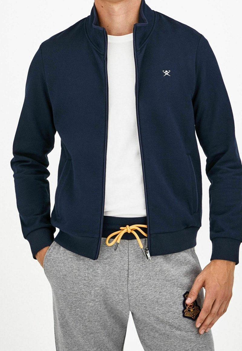 Homme portant une veste zippée bleu marine sur une chemise blanche, un pantalon de jogging gris avec un cordon jaune et un écusson brodé sur la jambe, mains dans les poches.