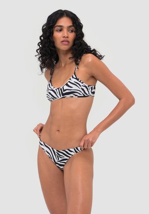 Kobieta o kręconych włosach, ubrana w czarno-biały bikini w wzór zebry, poprawiająca dolny element, patrząca z boku na jednolitym tle.