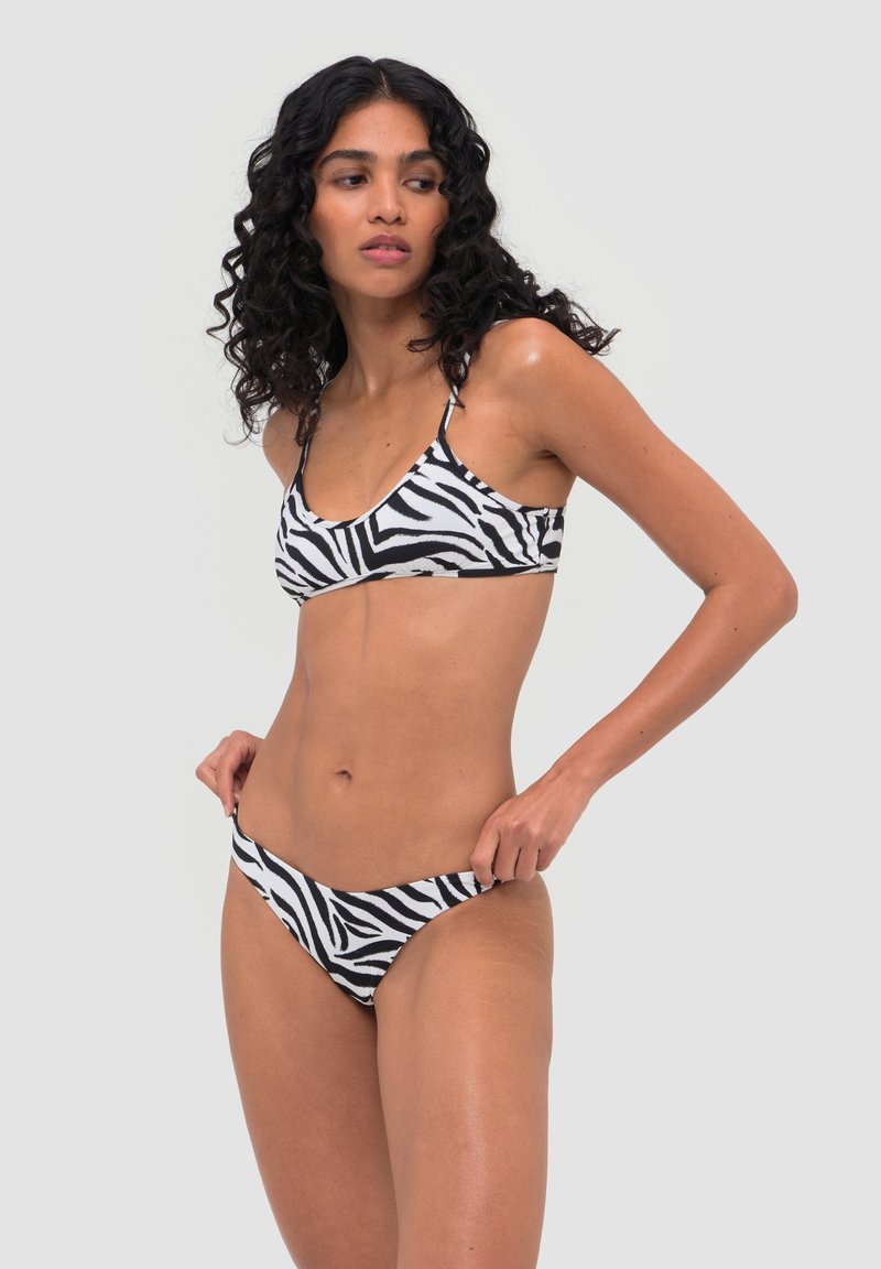 Femme aux cheveux bouclés portant un bikini à motif zèbre noir et blanc, ajustant la partie inférieure, regardant sur le côté devant un fond uni.