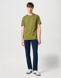 Camiseta de algodón verde oliva con mangas cortas, cuello redondo, pequeño detalle del logo en el pecho, combinada con jeans de color azul oscuro y zapatillas deportivas grises.