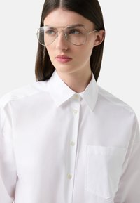 Witte blouse met een kraag, linkerschouderzak en een zachte textuur. Model draagt grote, dunne, ronde brillen. Schone uitstraling.
