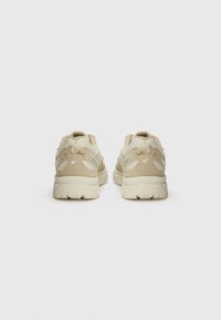 Beige athletic shoes with mesh and suede materials, lace-up design, cushioned sole, and subtle branding accents on the heel.