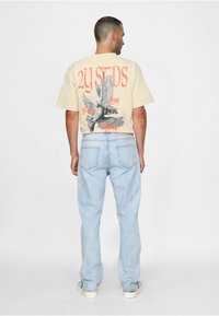 Beige t-shirt med en grafik av en duva och röd text på baksidan. Ljusblå jeans med raka ben och en avslappnad passform. Sneakers med krämfärgade sulor.