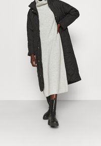 Cappotto lungo trapuntato nero con colletto alto, abbinato a un vestito lavorato a maglia grigio chiaro e stivali alla caviglia neri, che mostrano texture e un design a strati.
