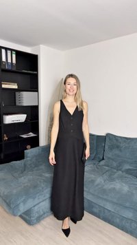 Melna bez piedurknēm kleita ar V-veida kakla izgriezumu, pogu aizdari un plūstošu dizainu. Valkāta ar melnām smailēm. Mīksta zila dīvāna fons.