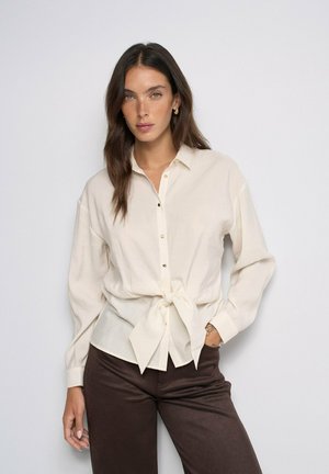 Mujer con cabello largo y castaño que lleva una blusa crema abotonada atada en la cintura y pantalones marrón oscuro sobre un fondo liso y claro.
