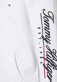 Sudadera blanca con texto bordado en negro "Tommy Hilfiger" y acentos rojos, gran bolsillo frontal y textura de tela suave.