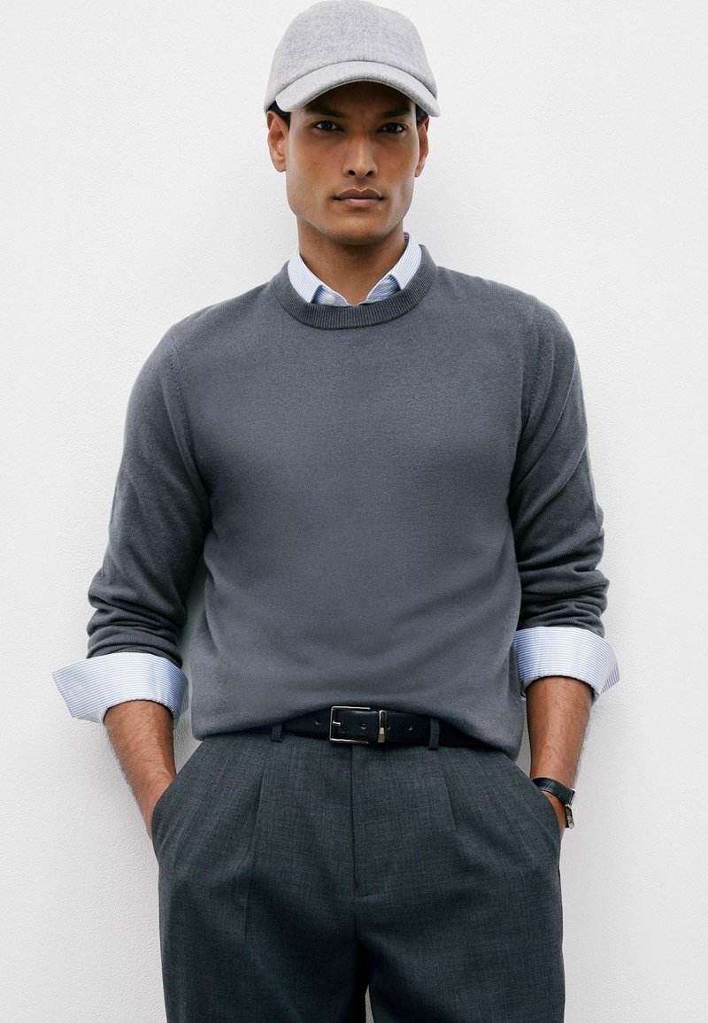Hombre con un suéter gris sobre una camisa rayada, pantalones grises, gorra gris, cinturón negro y reloj de pulsera, de pie con las manos en los bolsillos.