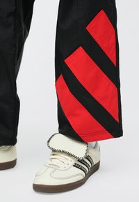 Čierne nohavice s červenými a čiernymi diagonálnymi pruhmi, doplnené bielymi teniskami s čiernymi pruhmi a polstrovaným jazykom.