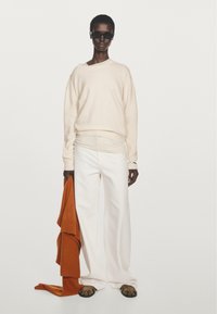 Cream sweater with a relaxed fit, white wide-leg trousers, and brown patterned sandals. Holding an orange knitted item. Black sunglasses worn.