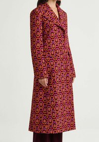Cappotto lungo con motivo geometrico nelle tonalità viola, arancione e rosa. Colletto con risvolto, chiusura con bottoni e tasche laterali. Tessuto dalla texture liscia.