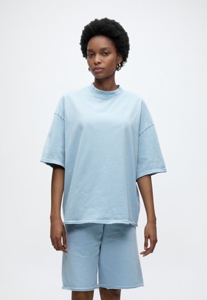 Žena s krátkymi kučeravými vlasmi, oblečená v svetlomodré nadrozmerné tričko s krátkym rukávom a zodpovedajúce krátke nohavice nad kolená, stojaca pred obyčajným bielym pozadím.