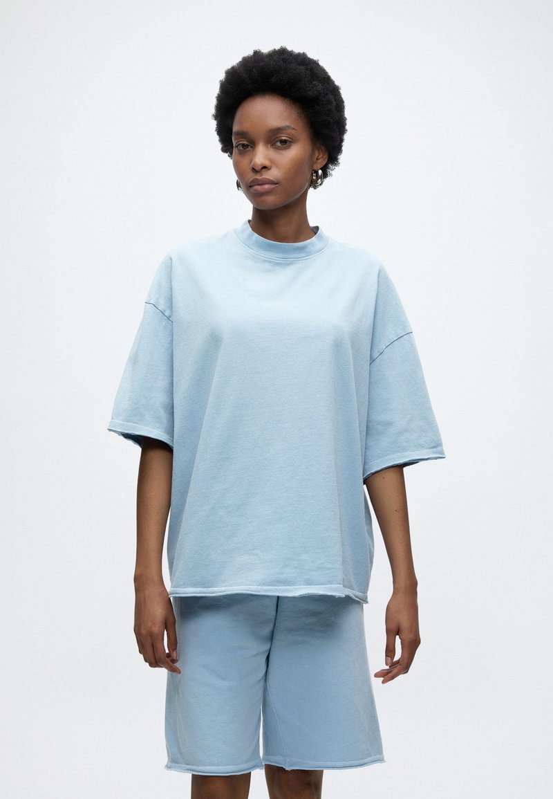 Γυναίκα με κοντά σγουρά μαλλιά φορώντας ανοιχτό μπλε φαρδιά κοντομάνικη μπλούζα και ταιριαστό σορτς μέχρι το γόνατο, στέκεται μπροστά σε απλό λευκό φόντο.