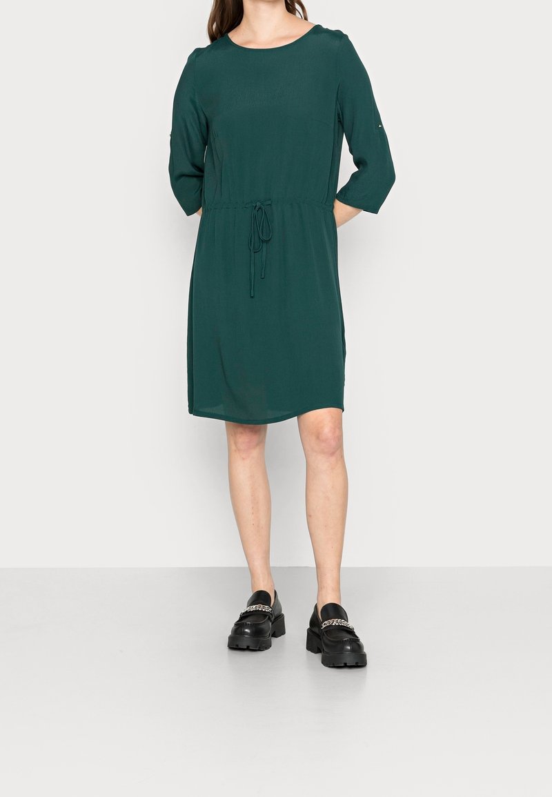 Femme portant une robe vert forêt mi-longue avec des manches trois-quarts et des mocassins noirs épais ornés de chaînes argentées.