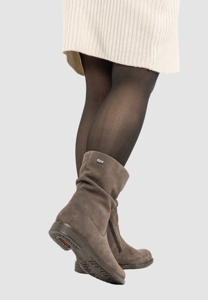Legs in black tights wearing beige suede mid-calf boots with side zipper and a cream ribbed knit skirt against a light background.