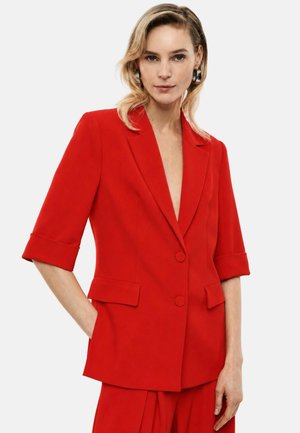 Donna con capelli biondi che indossa un blazer rosso brillante con le maniche arrotolate e pantaloni rossi abbinati su uno sfondo bianco semplice.