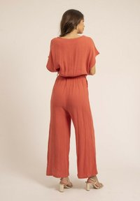 Combinaison rouge-orange en tissu léger et texturé. Coupe ample, jambes larges et taille élastique. Design simple et décontracté avec des manches courtes.