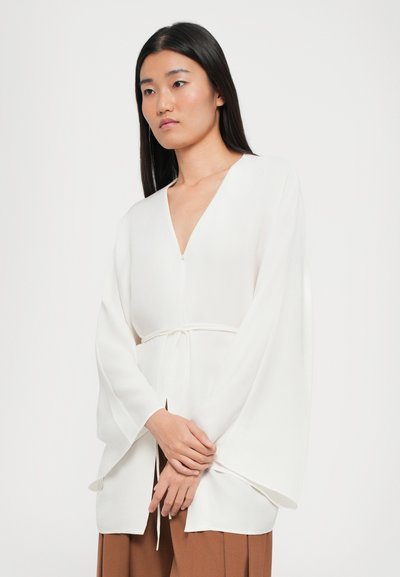 White, long-sleeve blouse with a V-neck and a tie at the waist; made of lightweight fabric with soft, smooth texture.