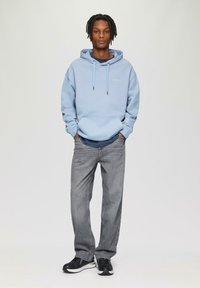 Lichtblauwe hoodie met een buidel, gedragen over een marineblauwe shirt, gecombineerd met grijze spijkerbroek en zwarte sneakers. Eenvoudige, casual outfit.