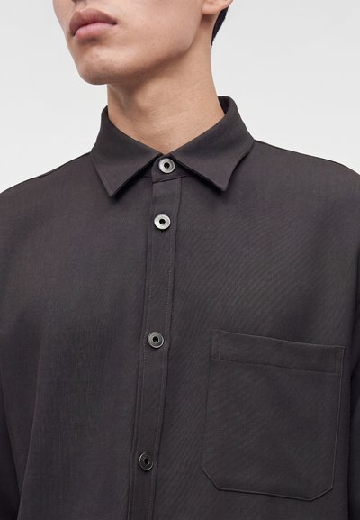 Personne portant une chemise sombre à boutons avec un col pointu et une poche poitrine, visible du cou à la moitié du torse sur un fond uni.