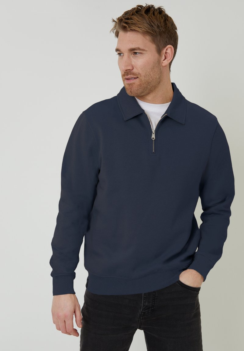 Marineblauer Pullover mit Kragen und Reißverschluss. Lange Ärmel und gerippte Bündchen. Getragen über einem weißen T-Shirt, kombiniert mit schwarzen Jeans.