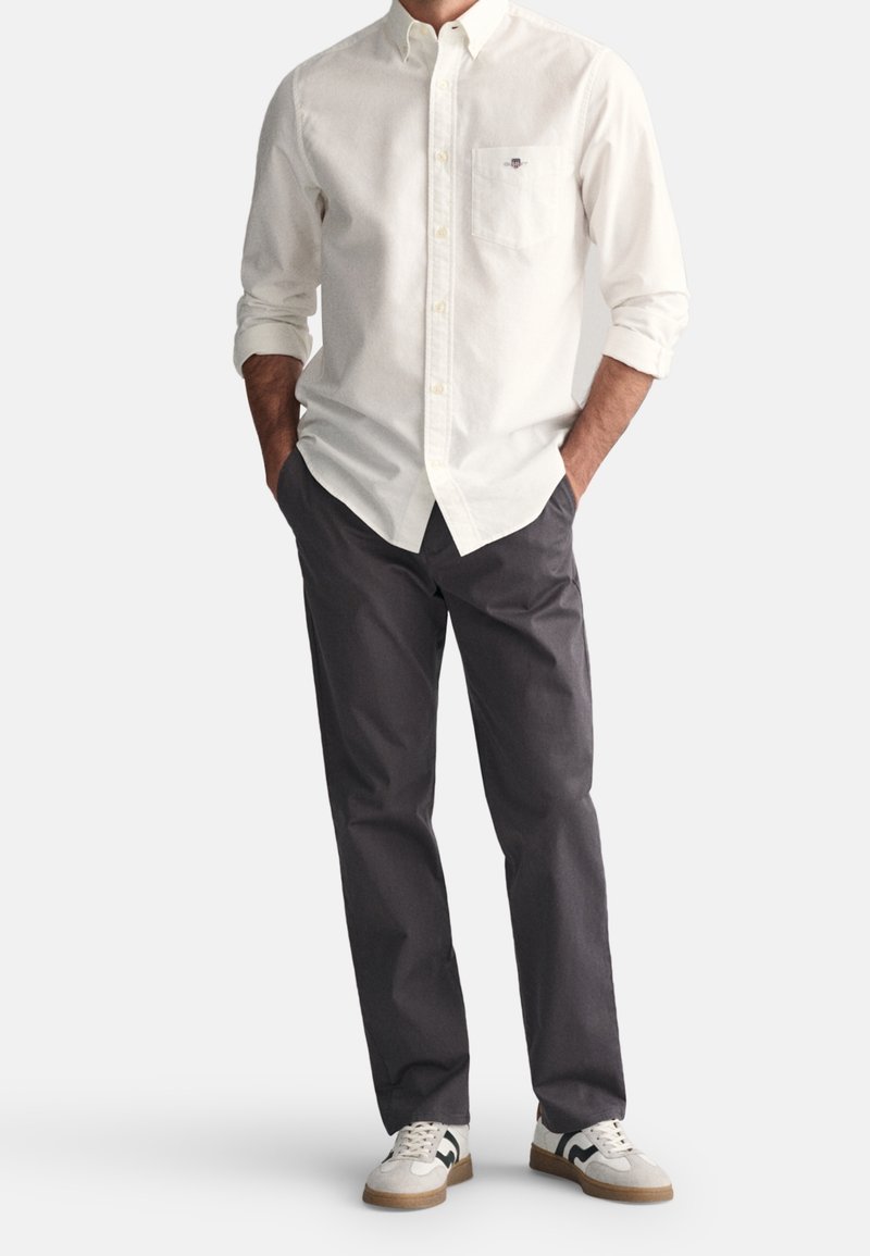 Homme portant une chemise blanche à boutons avec une poche poitrine, un pantalon gris foncé et des baskets blanches avec des détails noirs, debout les mains dans les poches.