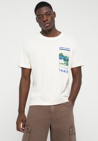 Beige bomulls T-skjorte med rund hals og korte ermer. Har et fargerikt lommeprint inspirert av Hokusai. Flatt stoff, avslappet passform.