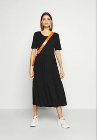 Vestido preto, até ao joelho, com mangas curtas, design em camadas e tecido suave. Usado com uma mala de ombro laranja brilhante e ténis brancos.