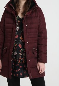 Giacca a vento bordeaux con cappuccio con zip, caratterizzata da una texture trapuntata, dettagli in argento e due tasche frontali. Indossata sopra un vestito floreale.