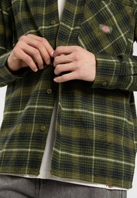 Een persoon past de knopen van een groen geruit flanellen overhemd aan, dat over een wit T-shirt is gedragen, met grijze broeken die gedeeltelijk zichtbaar zijn.