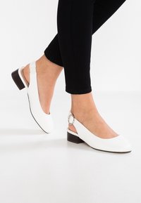 Ballerines blanches à talon slingback avec un bout rond, un petit talon marron et un accent en métal sur la bride de la cheville. Matériau lisse et design épuré.