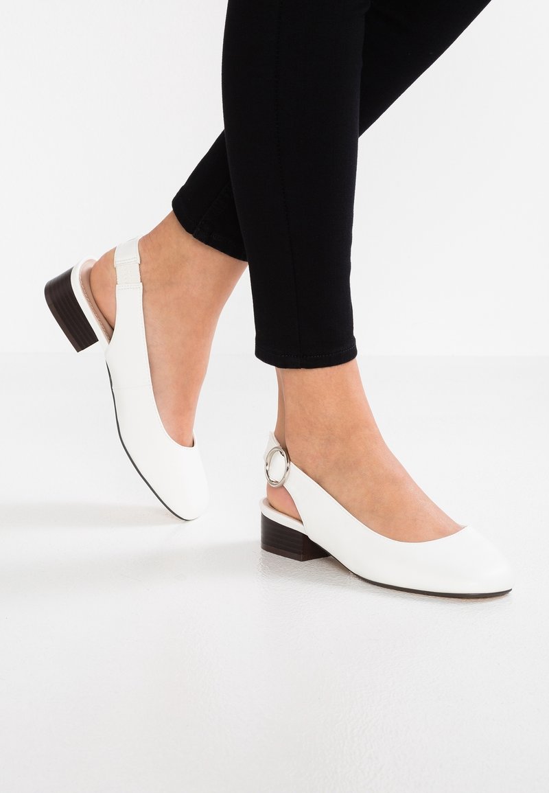 Ballerines blanches à talon slingback avec un bout rond, un petit talon marron et un accent en métal sur la bride de la cheville. Matériau lisse et design épuré.